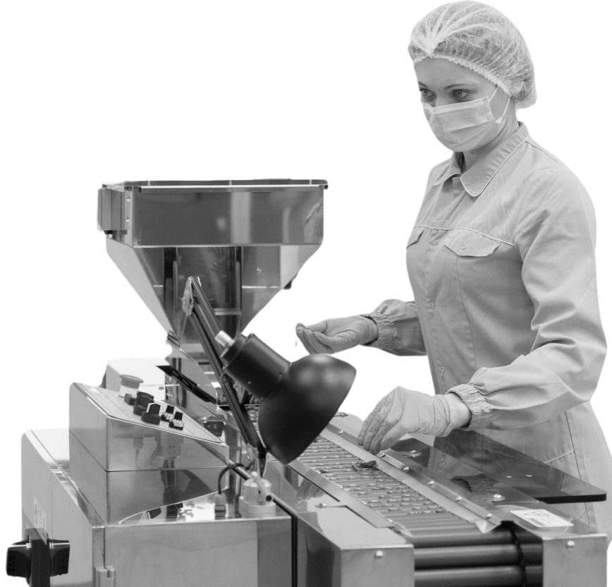 Black and white image of a female worker in a pharmaceutical or food production facility wearing a hairnet, face mask, and gloves while operating an industrial tablet or capsule filling machine.