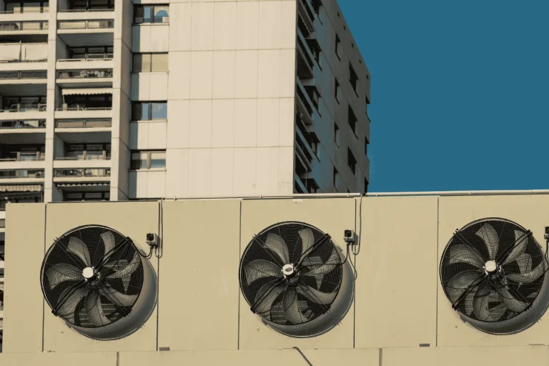 Three large industrial ventilation fans mounted on a rooftop with residential buildings in the background.